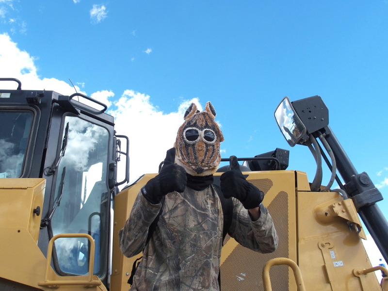 A protester in goggles and a 'chipmunk' full head mask gives a double thumbs up sign in front of a halted construction vehicle, against a blue sky with a few clouds.