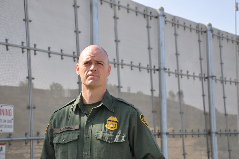 A US border patrol officer stands in front of a border wall.