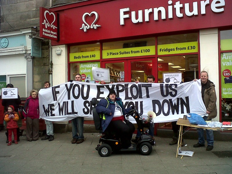 In front of a Heart Furniture store, protesters gather against workfare, of different ages and one in a wheelchair. A banner reads: If You Exploit Us We Will Shut You Down.