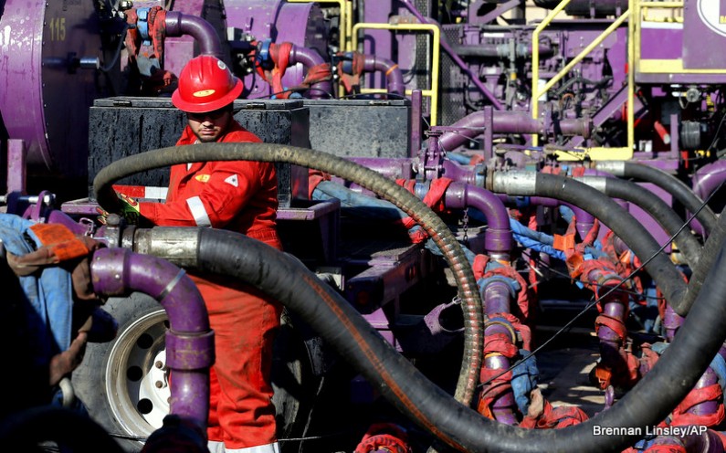 Surrounded by hoses and pipes, a worker in a jumpsuit and hard hat adjusts a hose.