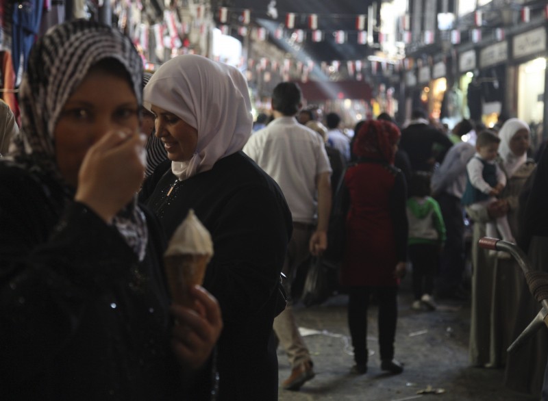 A woman in a hijab eats an ice cream cone. Behind her, others shop the bazaar, some also wearing head scarves.