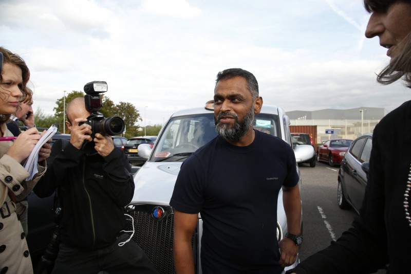 Moazzam Begg, wearing a dark t-shirt, speaks with reporters and photographers in a parking lot.