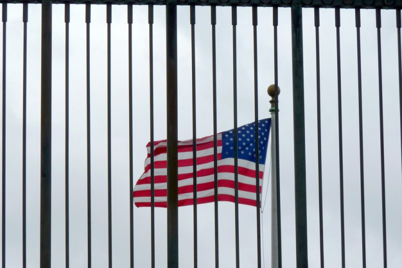 An American flag seen from behind a fence, so that it appears to be behind prison bars.