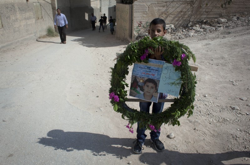 On a dusty street, a young boy carries a wreathe surrounding a memorial poster.