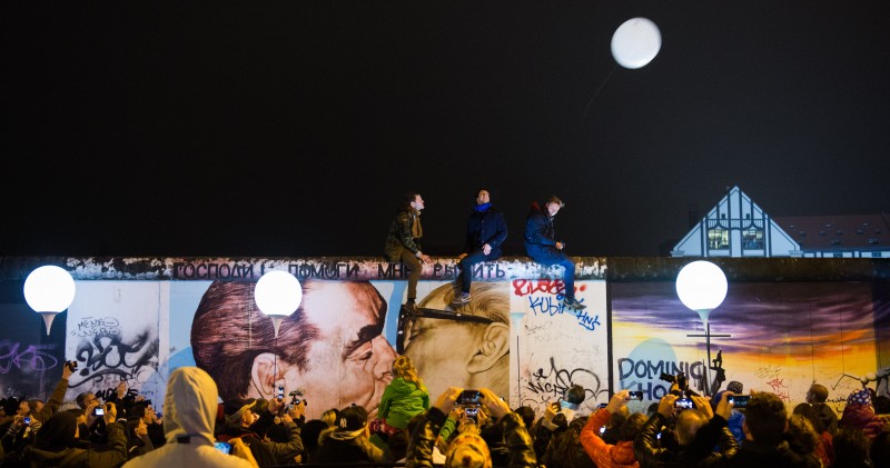 Lighted balloons rise past the remnants of the wall, covered in colorful graffiti. They are watched by a large crowd, three of whom have climbed atop the wall.