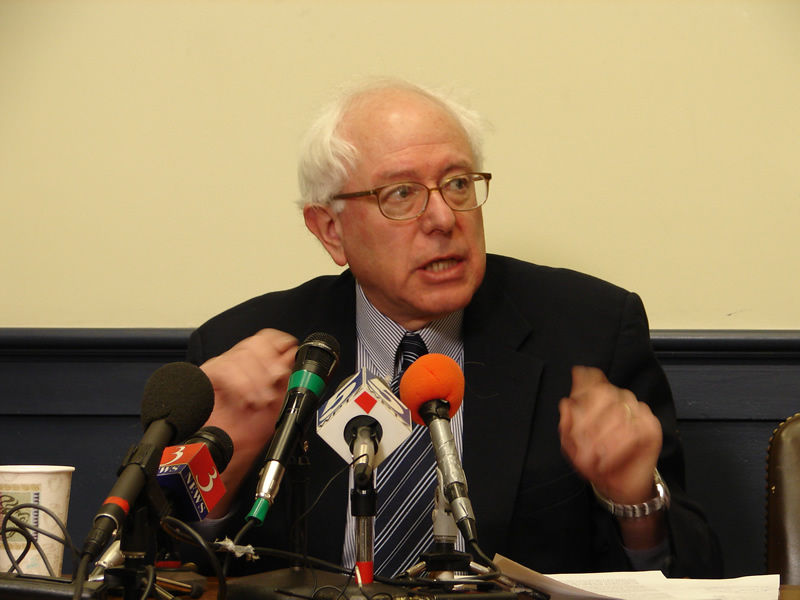 Bernie Sanders, wearing a suit, stands at a podium covered in microphones.