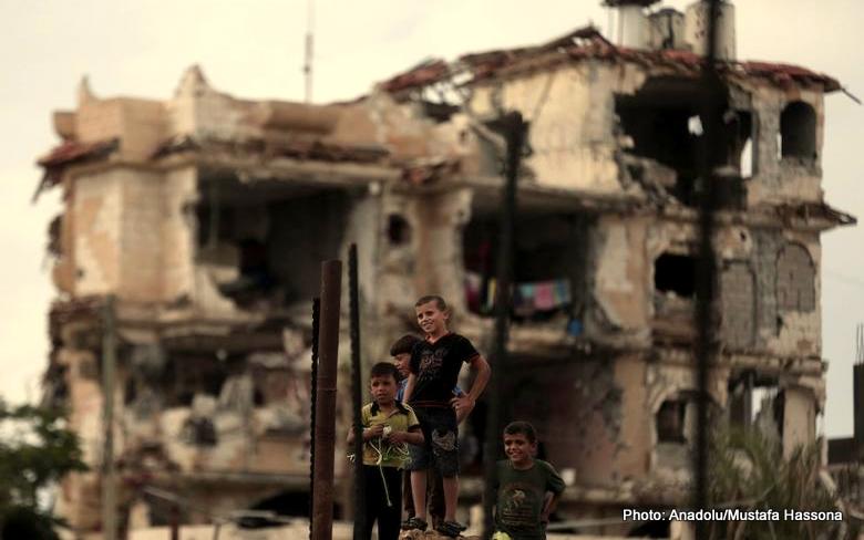 Three children sit in front of a pile of rubble which used to be housing.