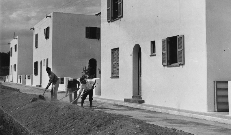 Three workers do road work with shovels in front of new, elegantly simple houses.