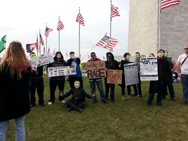 Masked activists hold signs for a diverse number of causes, arrayed in a row in front of the Monument.