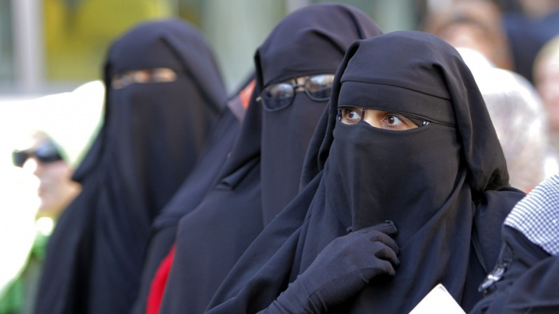 A line of three women in black niqab. One is wearing prescription glasses outside her veil.