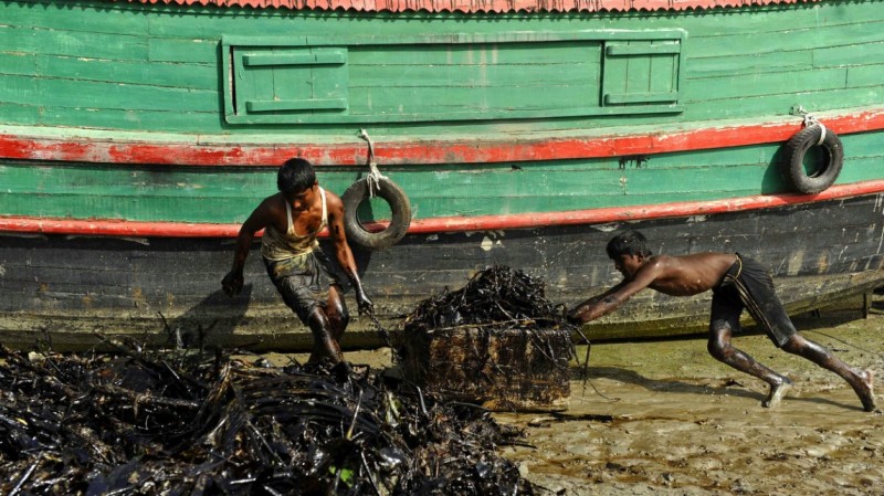 Two young men push heaps of oil slicked refuse along a beach beside the hull of a colorful boat.
