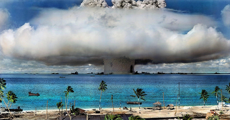 From the beach of a tropical island dotted with palm trees and huts, a massive mushroom cloud from a nuclear blast can be seen on the neighboring island.