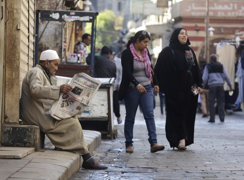 A man reads a newspaper on a stoop while two women approach on the street. 