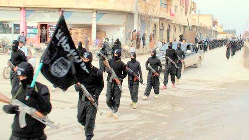 A row of black clad militants on parade in single file, faces covered in balaclava. The lead is holding the ISIS flag.