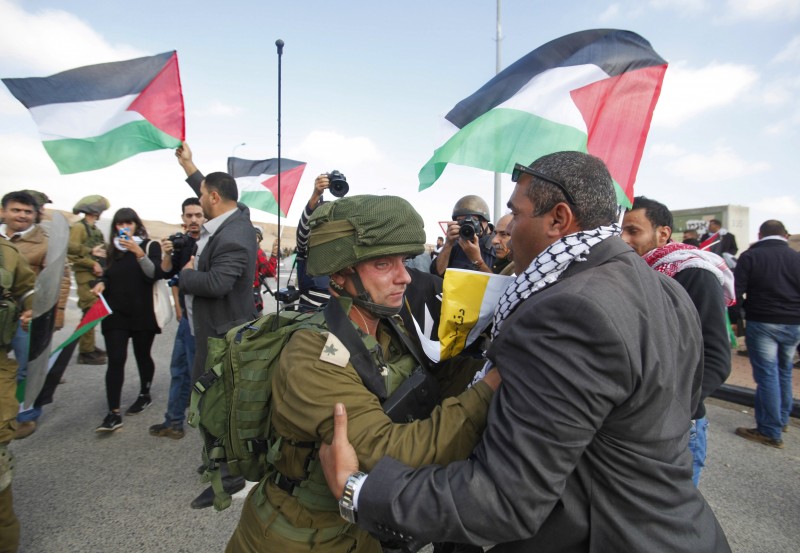 As the soldier and protester face off, other protesters waving Palestinian flags are seen in the background.