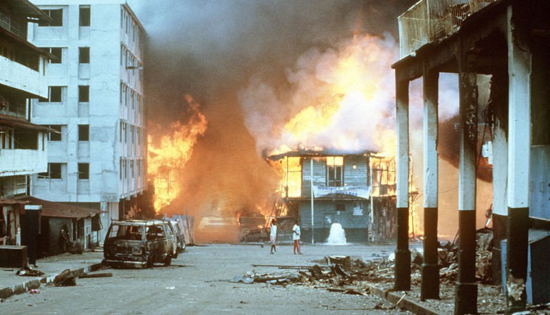 Barefoot civilians leave a burning building as flames roll into the sky. Nearby is a burnt out vehicle and other rubble.