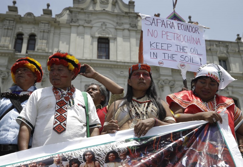 Indigenous activists in traditional clothes protest. One bilingual sign reads Keep The Oil In The Soil!