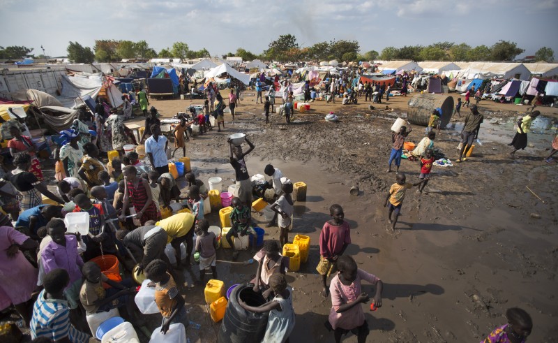 A huge crowd gathers on muddy, desolate ground around water containers with a refugee tent city in the distance.
