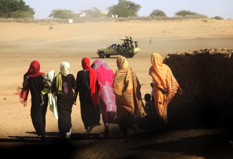 A row of women seen from behind, wearing colorful clothing and matching headscarves, walks into the desert. In the distance a military vehicle.