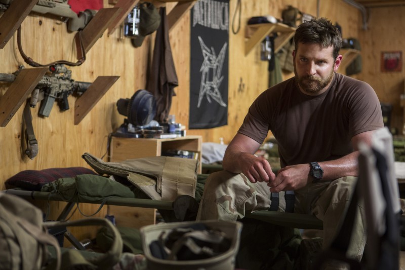 In this image released by Warner Bros. Pictures, Bradley Cooper appears in a scene from "American Sniper" as sniper Chris Kyle. He is sitting on a cot wearing military fatigues and a dark t-shirt. He is surrounded by military gear, with rifles mounted nearby on the wall.  (AP Photo/Warner Bros. Pictures, Keith Bernstein)