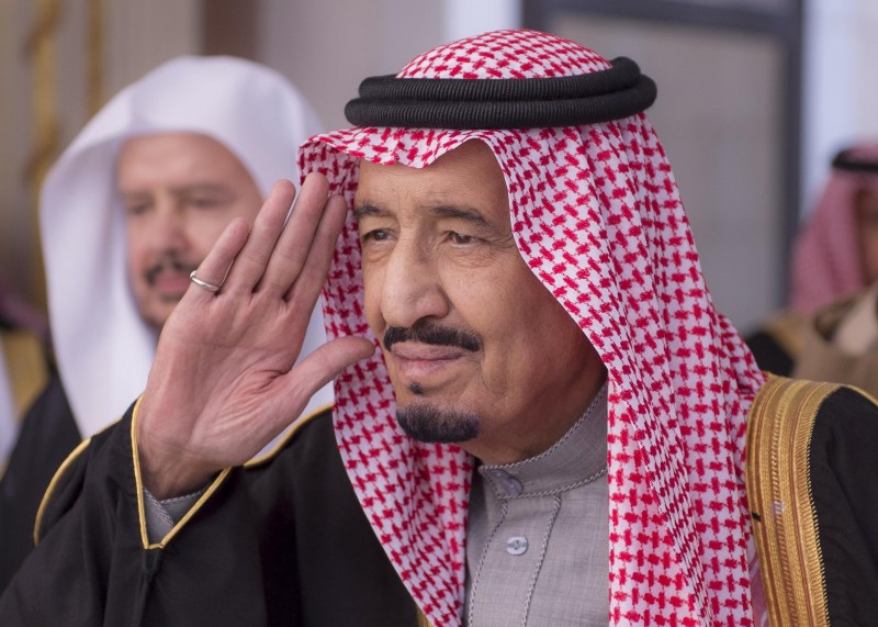 The prince makes a gesture like a salute, dressed in a white and red checked traditional keffiyeh.