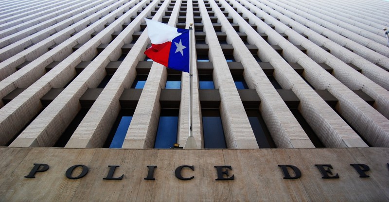 A look up at the facade of a tall office building, the words Police Dept are visible, with a Texas flag flying over them.