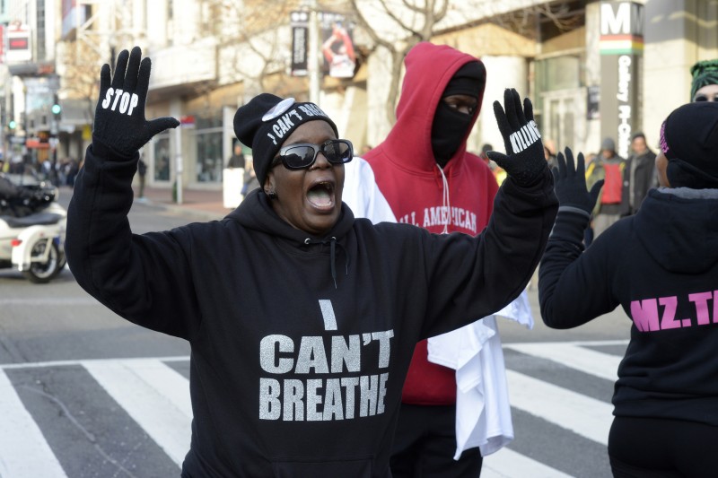 A protester has her hands up as she shouts. Her gloves read Stop Killing while her hat and sweatshirt read I Can't Breathe.