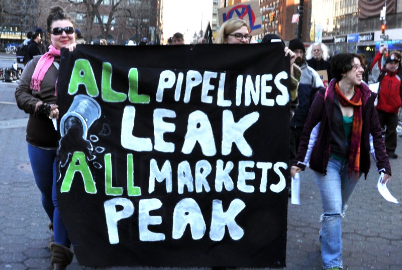 Two women march while carrying a banner that reads All Pipelines Leak All Markets Peak.