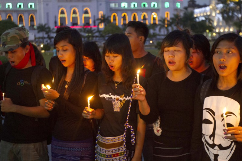 A row of activists holding candles in mourning for a protester, one wears a Guy Fawkes mask shirt.