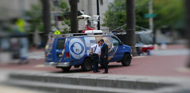 Two journalists speak near their satellite uplink van, labelled with the logo of a TV station.