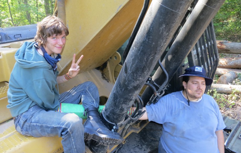 Shannon flashes a peace sign, seated atop the machinery while Ben stands nearby, both locked to the equipment.