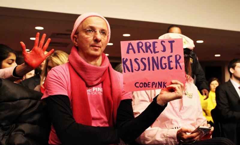 A codepink protester holds up a sign that reads Arrest Kissinger.