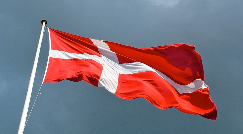Against an overcast sky, the flag of Denmark blows in a strong wind. Could other nations benefit by implementing Denmarks policies of ending inequality through high tax rates and a guaranteed social safety net? (Wikimedia / Per Palmkvist Knudsen)