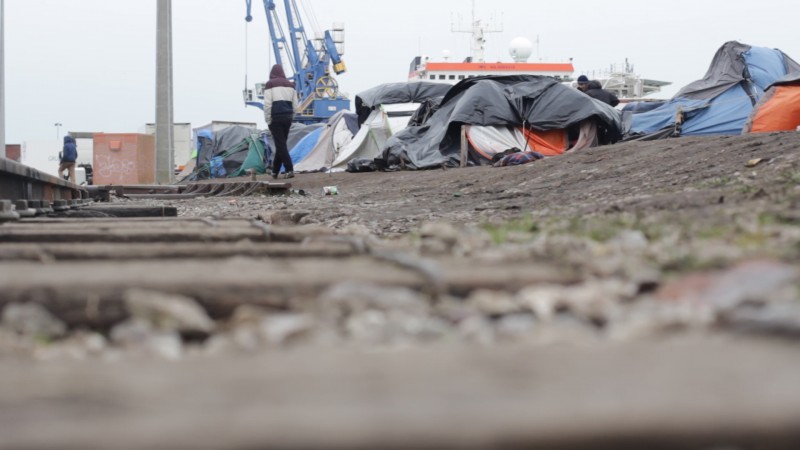 A man past a row of tarp-covered tents on a desolate patch of ground, with the cranes of the Port Of Calais, France visible in the distance. Hundreds of refugees from Syria's civil war ended up in this village while attempting to reach the United Kingdom. (Amel Guettatfi)