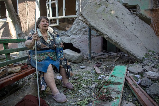 An older woman with a cane sits amid the rubble of a severely damaged stone home. International corporations are not the answer to rebuilding rural infrastructure destroyed by war. (Andrey Stenin)