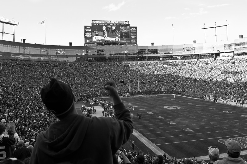 A Green Bay Packers fan stands in a crowded stadium with his fist raised in celebration, photographed on September 14, 2014. Despite the intense loyalty of many sports fans, most professional sports teams do little to benefit their local communities. (Flickr / Phil Roeder)