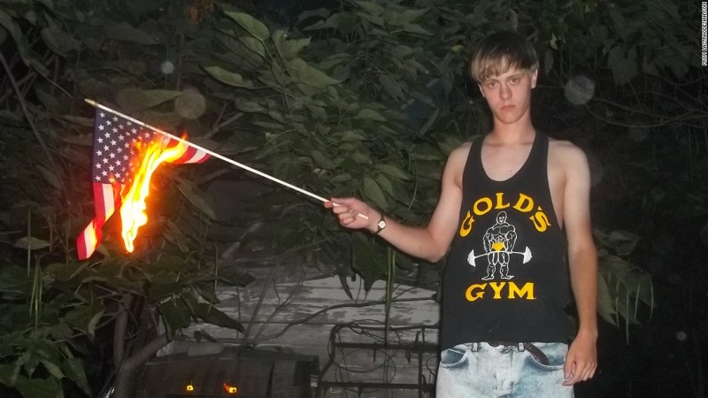 In this photo, Dylan Storm Roof, the accused killer of 9 black worshipers at a South Carolina church, butns an American flag outdoors while wearing a Gold's Gym tank top.  (Lastrhodesian.com)