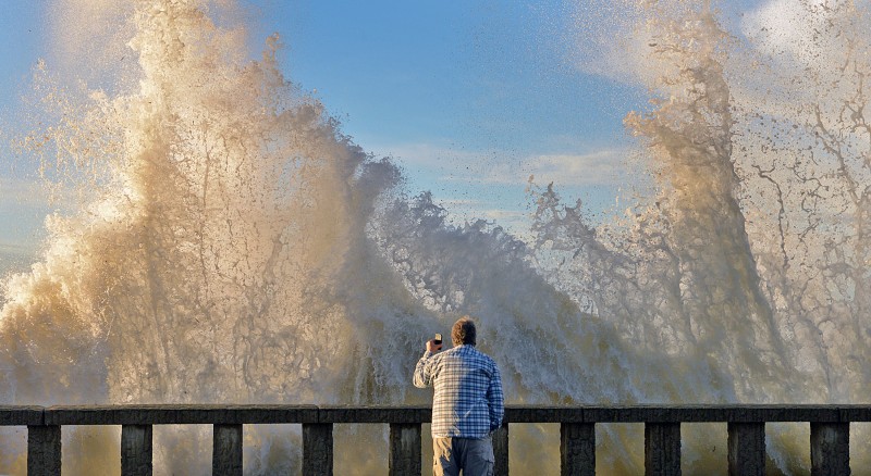 In a photo provided by Mike Eliason, Bo Sailor of Goleta, Caif. watches as high surf crashes into the wall and spills onto Channel Drive in Montecito, Calif. on Thursday, Jan. 7, 2016. The ocean-water-quality advisory issued Thursday came as the latest storms moved east after pummeling the region with heavy rainfall. Bacteria levels can increase significantly during and after rainstorms as contaminants in the runoff enter the ocean via storm drains, creeks and rivers. (Mike Eliason via AP)