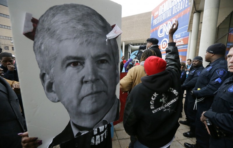 A protestor holds a sign with a photo of Michigan Gov. Rick Snyder before a Democratic presidential primary debate at the University of Michigan-Flint, Sunday, March 6, 2016, in Flint, Mich. (AP Photo/Carlos Osorio)