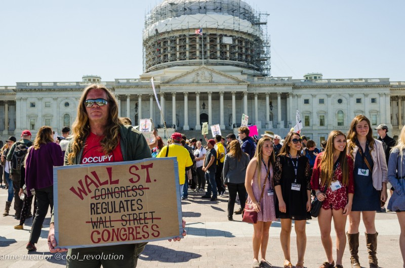 In front of a large rally gathered at the U.S. Capitol, an activist holds a sign which reads "Wall Street Regulates Congress." April 13, 2016. (Flickr / cool revolution)