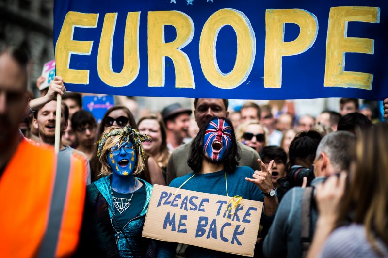 July 2, 2016. Under a "Europe" banner at a protest against the Brexit vote, two activists wear UK flag face paint. One wears a sign around his neck which reads "Please take me back." (Flickr / Garon S)