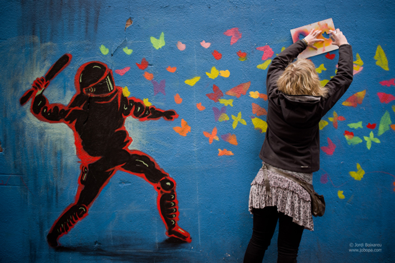 In the streets of El Raval neighborhood in Barcelona, a graffiti artist creates a mural, using a stencil, which shows a riot cop attempting to beat a swarm of butterflies. February 21, 2015. (Flickr / Jordi Boixareu)