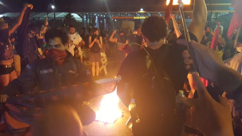 In this July 27, 2016 photograph, masked anti-capitalist activists burn a U.S. flag near the Democratic National Convention in Philadelphia. (Kit O’Connell)