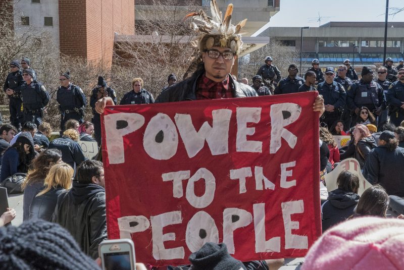 At an anti-Trump protest at the University of Wisconsin, a Native American activist in a feather headpiece holds a "Power To The People" banner. January 1, 2014. (Flickr / Joe Brusky)