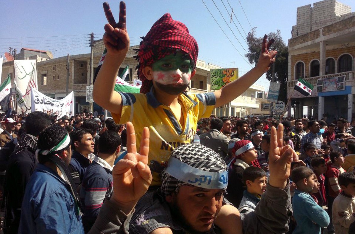 In this citizen journalism image taken on Monday March 26, 2012 and provided by Edlib News Network ENN, an anti-Syrian regime protester carries a boy on his shoulder with the Syrian revolution flag painted on his face, gesture during a demonstration in Binish town in Idlib province, northern Syria. (AP Photo/Edlib News Network ENN)