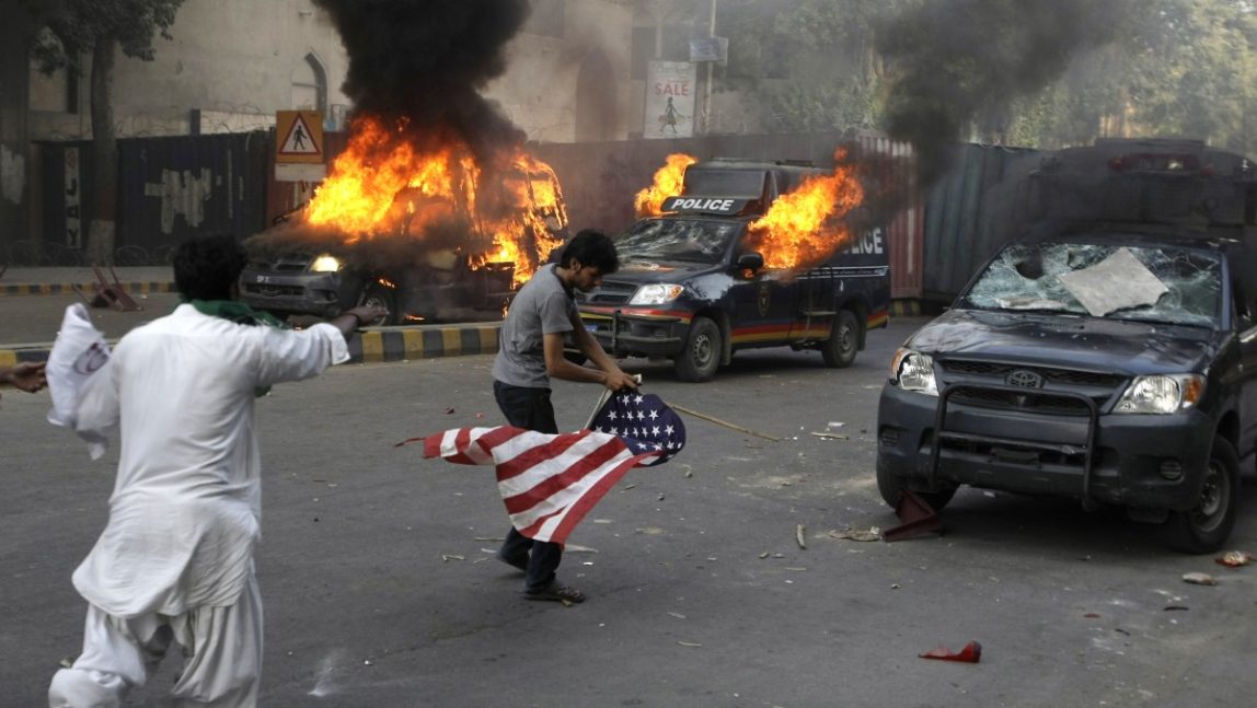 A protester carries a representation of a U.S. flag as police vehicles burn in Karachi, Pakistan on Friday, Sept 21, 2012. (AP Photo/Fareed Khan)