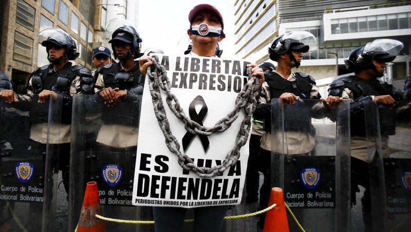 A woman with her mouth covered stands before a line of National Bolivarian Police preventing protesters from reaching the national intelligence agency in Caracas, Venezuela, Tuesday, Feb. 11, 2014. Her sign reads in Spanish "Freedom of expression. It's yours. Defend it." (AP Photo/Alejandro Cegarra)