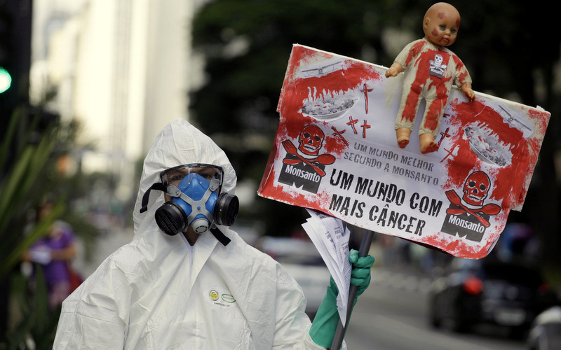 A protester wearing a protective suit and mask holds up a sign toward passing cars that reads in Portuguese "A better world according to Monsanto is a world with more cancer" in Sao Paulo, Brazil, Saturday, May 25, 2013. (AP/Nelson Antoine)