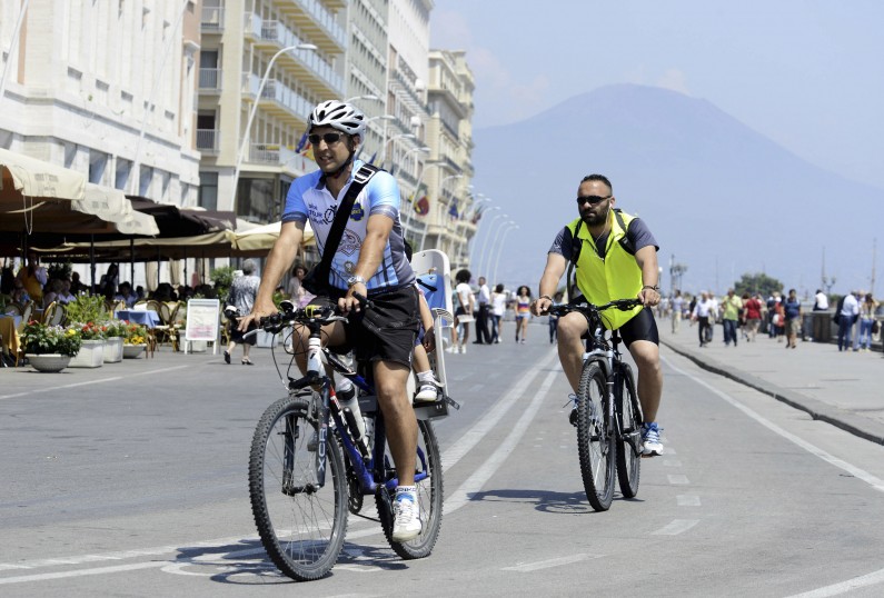 In this Friday, June 13, 2014 photo, Luca Simeone, founder of bike-sharing start up called "bike tour Napoli," left, pedals along a cyclists' path in Naples, Italy. In the background is Mt. Vesuvius. Three years ago Naples seafront was an urban highway, noisy and smoggy, jammed with car traffic, while smelly trash erupted from garbage bins along streets and alleys. Urban cyclers were regarded as eco-fundamentalists. Three years later, Naples has a new mayor, clean streets, a wide pedestrian beachfront and a 20-kilometer (20-mile) cycling lane overlooking a beautiful bay.  (AP Photo/Salvatore Laporta)