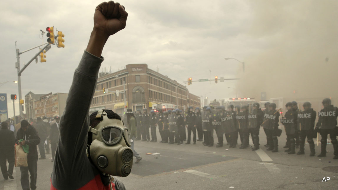 A demonstrator raises his fist as police stand in formation as a store burns, Monday, April 27, 2015, during unrest following the funeral of Freddie Gray in Baltimore.